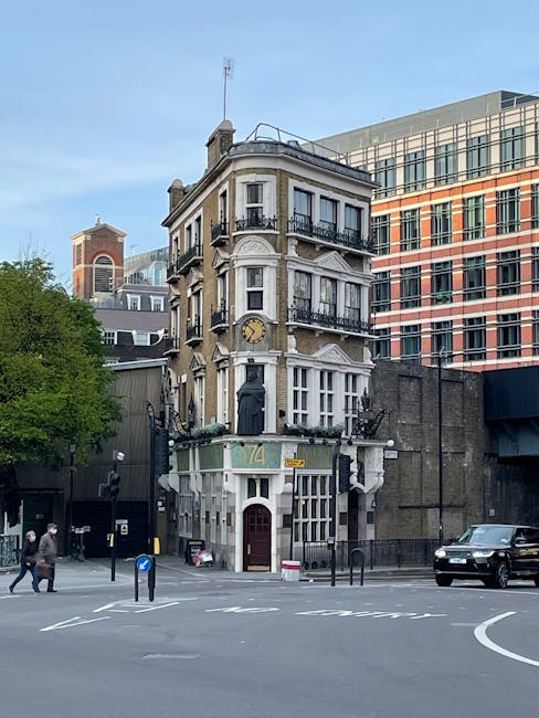 A three-storey historic building located at a street corner in Blackfriars during daytime, with classical architectural details, black wrought iron balconies, and a large clock on the facade. The ground floor features a white and green painted exterior with a red door and the number 174. Surrounding the building are modern office structures with large glass windows, and a tree on the left side. In the foreground, there is a paved crosswalk, a few pedestrians, and a vehicle parked nearby. This scene reflects urban settings where home relocation and furniture transport services by Man and Van Blackfriars may be required during house removals or moving logistics, with the building potentially serving as the subject of a moving or packing process involving loading furniture or boxes near the property entrance.