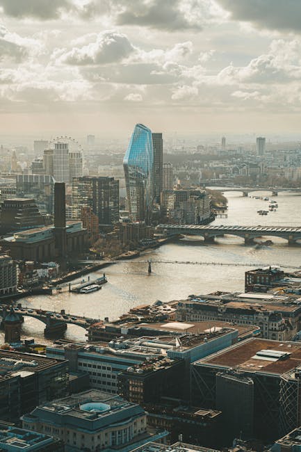 An aerial view of a cityscape featuring a river running through the urban area with multiple bridges crossing over it. Tall modern buildings, including a distinctive blue-glass skyscraper, dominate the skyline under a partly cloudy sky with soft sunlight illuminating the scene. In the foreground, rooftops of various commercial and industrial structures are visible, while waterways and boats can be seen flowing past. This image captures a bustling city environment that is suitable for illustrating urban home relocations and professional removals services offered by Man and Van Blackfriars, highlighting the logistical environment involved in furniture transport and moving processes within a major city.
