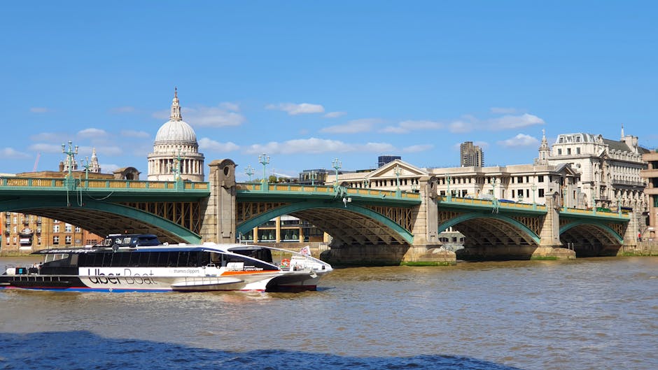 A white and black Uber Boat ferry is navigating along the River Thames beneath the historic Blackfriars Bridge in London, with the dome of St. Paul's Cathedral visible in the background. The river's water reflects the clear blue sky with scattered clouds. On the riverbank, there are several historic multi-storey buildings, and the bridge features a green upper structure supported by stone columns, with street lamps lining its edges. In the foreground, the ferry appears to be in the process of docking or departure, and the scene captures an urban environment typical of central London. This outdoor setting is part of a snapshot relevant to house removals and logistics, illustrating the concept of transport and access considerations near the Blackfriars area for moving services. Regular operational activities like vessel navigation are visible, underscoring the importance of awareness around river access for home relocation projects, as managed by [COMPANY_NAME].