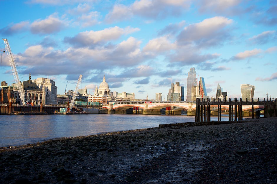 A wide view of the London city skyline taken from the south bank of the River Thames during daylight, showcasing notable buildings such as St. Paul's Cathedral and the modern skyscrapers of the financial district. The river in the foreground has a pebbled shoreline with some wooden pilings and a stone wall along the edge. The scene includes a partly cloudy sky with blue patches visible. In the middle ground, a bridge spans the river, supporting vehicular traffic. On the left side of the image, construction cranes and a historic building with a classical dome are visible. The overall setting suggests an urban environment with ongoing building development, relevant to house removals or furniture transport activities that might involve moving across such a cityscape. Man and Van Blackfriars occasionally conducts home relocation and furniture transport services in this area, although these activities are not visible in this particular image.