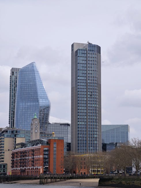 A cityscape featuring modern skyscrapers, including a distinctive curved glass building and a tall rectangular office tower, situated behind a row of brick and concrete buildings with traditional architecture. In the foreground, leafless trees line the waterfront promenade, which is empty of people. The cloudy sky overhead reflects typical urban weather. This scene is captured during daylight, illustrating the urban environment around Blackfriars Station, relevant to home relocation and moving services by Man and Van Blackfriars, who provide professional removals and furniture transport in the area.