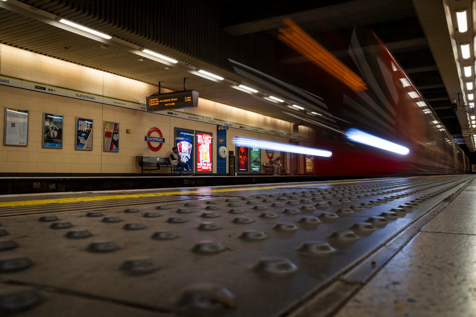 A long-exposure photograph of a train arriving or departing at Blackfriars underground station, with the movement creating a blurred red and white streak across the platform. The station’s beige tiled walls are decorated with posters and signs, including the recognizable London Underground roundel. There are illuminated screens displaying train times hanging from the ceiling, and a few commuters sitting on benches near the wall. The platform’s tactile paving and safety yellow line are visible in the foreground, and the scene is lit by overhead fluorescent lights. This setting captures the busy atmosphere of home relocation logistics involving furniture transport and packing in the context of house removals, with the station serving as a key transit point for moving services like those provided by Man and Van Blackfriars.