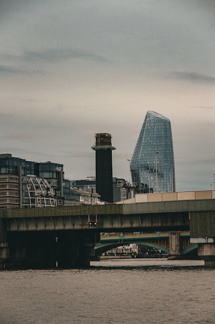 A view of a cityscape featuring modern buildings and a bridge over a river, with one building displaying a distinctive slanted glass facade, typical of urban architecture in Blackfriars. The scene appears overcast, with grey skies casting diffuse light across the structures. The bridge, made of concrete and steel, spans the river and supports vehicular traffic, with visible roadways and supports beneath. In the foreground, the river surface is calm, reflecting parts of the bridge. This setting highlights an environment where house removals and furniture transport may involve navigating dense urban infrastructure, including bridges and river crossings, as part of home relocation or moving services offered by companies like Man and Van Blackfriars.