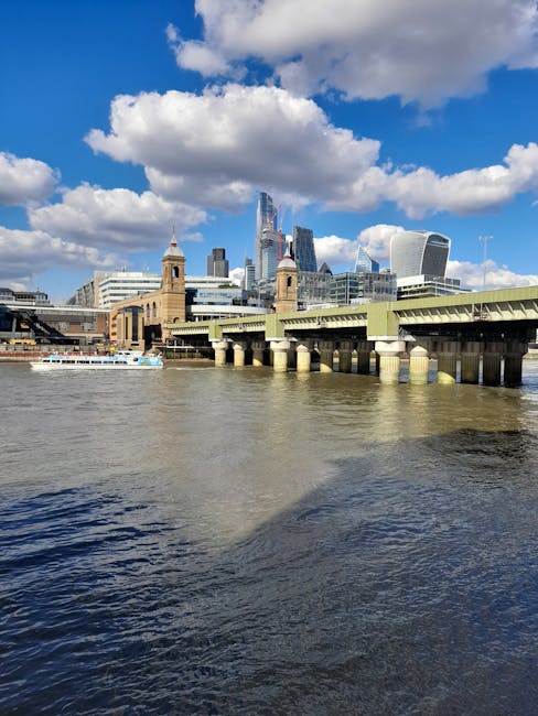 A view of the River Thames in London during daytime, with the Tower Bridge in the foreground and the modern skyscrapers of the City of London in the background under a partly cloudy sky. The bridge's stone towers and green suspension elements are visible along with a boat on the water. This urban landscape highlights the contrast between historic and contemporary architecture, relevant to property relocations and moving logistics in central London, as managed by Man and Van Blackfriars, who provide comprehensive removals services including home relocation, furniture transport, packing, and loading processes for properties near or within the Blackfriars area.
