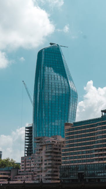 A view of a cityscape featuring modern buildings and a bridge over a river, with one building displaying a distinctive slanted glass facade, typical of urban architecture in Blackfriars. The scene appears overcast, with grey skies casting diffuse light across the structures. The bridge, made of concrete and steel, spans the river and supports vehicular traffic, with visible roadways and supports beneath. In the foreground, the river surface is calm, reflecting parts of the bridge. This setting highlights an environment where house removals and furniture transport may involve navigating dense urban infrastructure, including bridges and river crossings, as part of home relocation or moving services offered by companies like Man and Van Blackfriars.