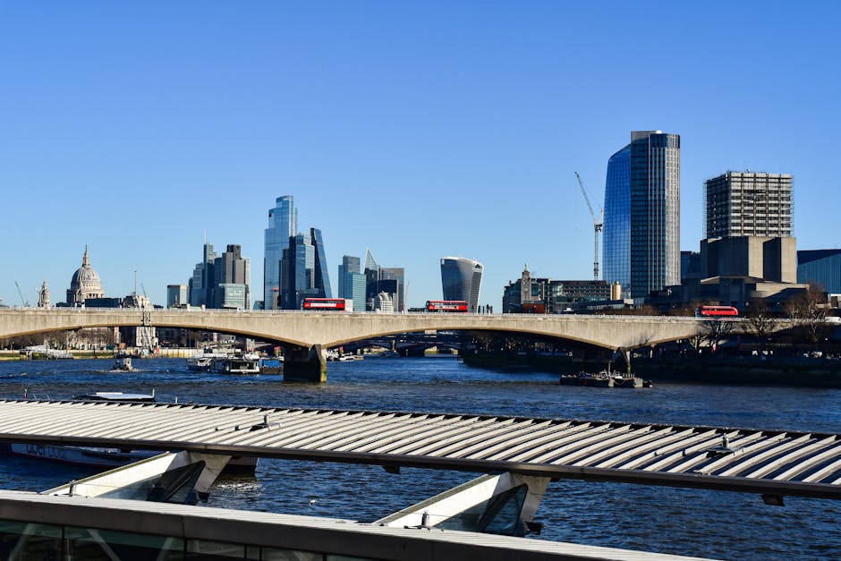 A view of London's modern skyline featuring tall office buildings and skyscrapers, with the historic St. Paul's Cathedral visible on the left side of the image. A concrete bridge spans the River Thames in the foreground, with several red double-decker buses traveling across it. Below the bridge, there are watercraft on the river, and part of a glass and metal structure is visible at the bottom of the image. The scene is captured during the day with clear blue skies, and the sunlight illuminates the buildings, casting gentle reflections on the water. This image highlights urban infrastructure and cityscape elements relevant to house removals and relocation logistics, such as transportation routes and building environments, as considered by companies like Man and Van Blackfriars when managing furniture transport and packing during home relocations.
