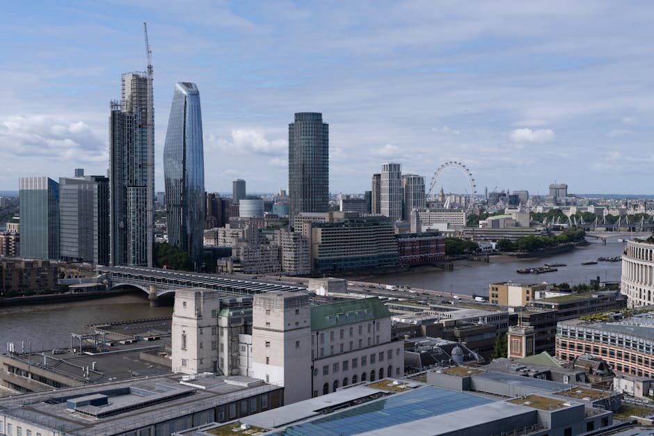 A wide view of London's skyline showing modern skyscrapers, including a diverse range of tall glass buildings and high-rise structures, with the River Thames flowing through the foreground. Several bridges cross the river, and boats are visible on the water. In the lower part of the image, rooftops and early-stage construction sites are seen, along with a white historic building closer to the camera. The scene is captured during daylight with partly cloudy skies, providing natural light that highlights the urban landscape. This image reflects the busy nature of city centre relocations, such as those managed by Man and Van Blackfriars, with the city's mix of commercial and historic architecture supporting efficient home and furniture transport services across central London.
