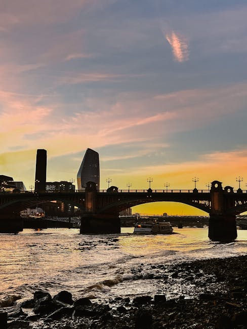 A cityscape at sunset shows a river with dark, rocky banks in the foreground, along with small boats on the water. A historic brick bridge spans the river, with arches visible beneath the roadway, supporting a line of streetlights and decorative elements. Behind the bridge, the modern skyline features several tall buildings, including a distinctive cylindrical tower and a slanted skyscraper, set against a colorful sky with hues of pink, orange, and blue. The overall scene captures an urban waterfront area, relevant to house removals and city-based relocation services provided by Man and Van Blackfriars, illustrating the type of environment where professional removals may operate in central London.
