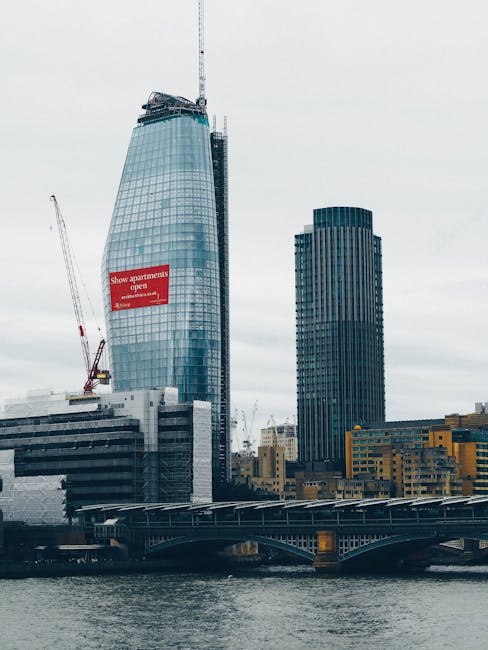 A tall, modern glass skyscraper under construction stands prominently in the image, with a construction crane attached to its upper section and a red banner advertising apartments displayed on its facade. Next to it is a dark, rectangular office building with a grid of windows, set against an overcast sky. In the foreground, a river runs beneath a bridge with a metal railing and supporting arches, with calm water reflecting the buildings above. The scene captures a cityscape in central London, with ongoing building work indicative of urban development. This environment, characterized by high-rise structures and waterway access, relates to house removals and relocation logistics, especially when planning furniture transport or moving services in busy urban areas, as provided by companies like Man and Van Blackfriars.