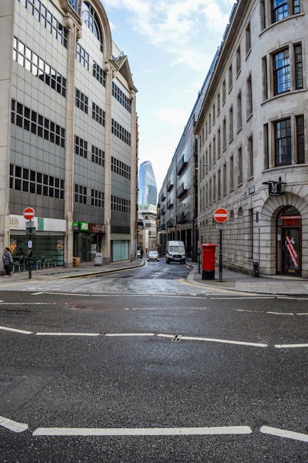 A street scene in the Blackfriars area showing a narrow city road flanked by tall, multi-storey office buildings constructed from light-colored stone and dark glass panels. In the foreground, the wet pavement features white dashed and solid lines, along with  road markings indicating traffic directions. Two red and white 'no entry' signs are mounted on poles on either side of the road. A white van is parked on the right side near a curb, adjacent to a red British telephone booth. On the left sidewalk, a person dressed in dark clothing is standing beside a small black bollard, near other street furniture and a shopfront with illuminated signage. The background reveals additional modern office buildings with reflective glass facades and a partly cloudy sky overhead, suggesting an active urban environment suitable for home relocation or furniture transport operations, as managed by Man and Van Blackfriars during a residential move or commercial removal process.