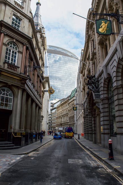 A street scene in the Blackfriars area showing a narrow city road flanked by tall, multi-storey office buildings constructed from light-colored stone and dark glass panels. In the foreground, the wet pavement features white dashed and solid lines, along with  road markings indicating traffic directions. Two red and white 'no entry' signs are mounted on poles on either side of the road. A white van is parked on the right side near a curb, adjacent to a red British telephone booth. On the left sidewalk, a person dressed in dark clothing is standing beside a small black bollard, near other street furniture and a shopfront with illuminated signage. The background reveals additional modern office buildings with reflective glass facades and a partly cloudy sky overhead, suggesting an active urban environment suitable for home relocation or furniture transport operations, as managed by Man and Van Blackfriars during a residential move or commercial removal process.