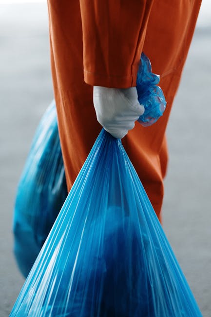 A person wearing orange protective overalls and white gloves holding a blue plastic bag filled with waste, standing on a paved area. In the background, a moving van or vehicle is partially visible behind the individual. The scene suggests the process of waste collection or disposal during home relocation or house clearance, with the focus on handling rubbish materials safely and efficiently, as part of professional removals services. The image emphasizes the careful handling of waste, appropriate packaging materials, and the environmental aspect of waste management in a moving or clearance context, aligned with the services offered by Man and Van Blackfriars.