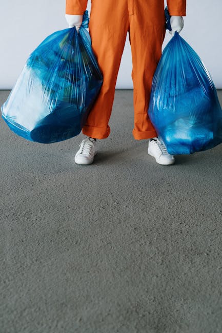 A person standing indoors on a grey carpet floor, wearing orange pants, white sneakers, and grey gloves, holding two large blue plastic garbage bags filled with household waste or unwanted items. The individual is positioned in front of a plain light-colored wall, with the bags hanging at each side, suggesting the process of home relocation or preparing for rubbish removal. Visible textures include the smooth plastic of the garbage bags, the fabric of the clothing, and the carpet surface underneath. This scene depicts the loading stage of a house clearance, typical of moving or disposal services provided by companies like Man and Van Blackfriars, situated in a room ready for waste collection or disposal during a house removal or packing procedure.