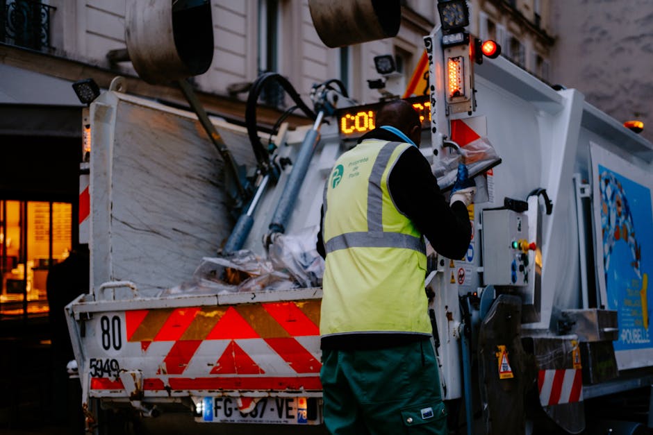 A person wearing orange protective overalls and white gloves holding a blue plastic bag filled with waste, standing on a paved area. In the background, a moving van or vehicle is partially visible behind the individual. The scene suggests the process of waste collection or disposal during home relocation or house clearance, with the focus on handling rubbish materials safely and efficiently, as part of professional removals services. The image emphasizes the careful handling of waste, appropriate packaging materials, and the environmental aspect of waste management in a moving or clearance context, aligned with the services offered by Man and Van Blackfriars.