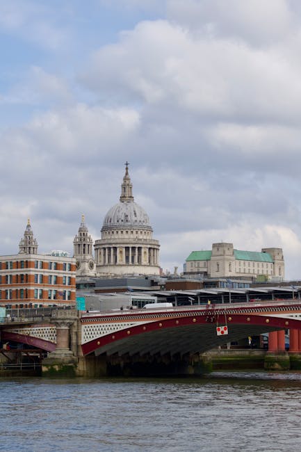 A view of St. Paul's Cathedral in London with its prominent domed roof and ornate architectural details, seen from the riverbank. In the foreground, a red bridge spans the river, supported by stone piers, with patches of water visible beneath. To the left, part of a modern brick building with large windows is visible, contrasting with historic structures. The sky above is partly cloudy with patches of blue, and the scene captures a typical daytime setting. This image reflects urban scenery relevant to house removals and furniture transport services, such as those offered by Man and Van Blackfriars, highlighting the importance of planning logistics for home relocation and loading processes in London.