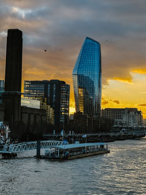 A cityscape featuring modern high-rise buildings with glass facades, including a distinctive curved structure and a tall rectangular tower, set against a cloudy sky. In the foreground, there are leafless trees along a paved walkway, with a low railing separating the area from the water. This urban environment reflects contemporary architecture typical of a professional environment where house removals and moving services, such as those offered by Man and Van Blackfriars, may operate during home relocation or furniture transport processes.